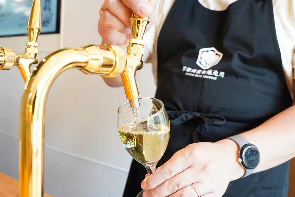 黒いエプロンを着た人が、金色に輝くサーバーから透明なグラスに黄金色の液体を注いでいます。エプロンには「京都蜂蜜醸造所」のロゴが見え、蜂蜜酒(ミード)を試飲または提供している様子です。