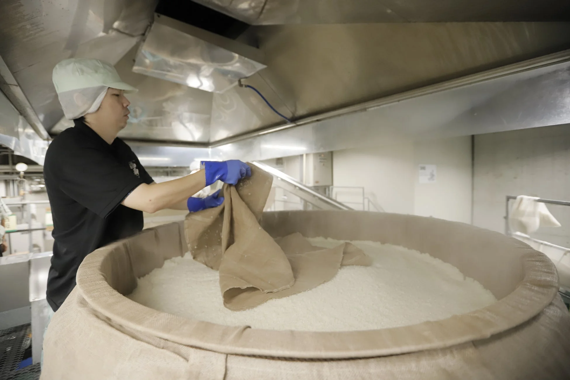 酒造工場で、作業員が大きな桶に盛られた蒸米や麹のような白い粒状のものを布で覆う作業をしている様子。