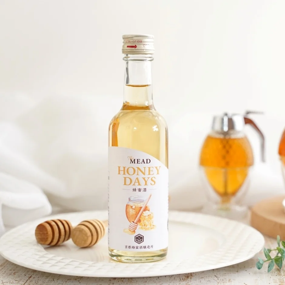 白い背景のテーブルに置かれた白い皿の上に、蜂蜜酒「HONEY DAYS」のボトルが中央に配置されています。ボトルの手前には木製のハニーディッパーが2本あり、背景にはぼかされたハニーディスペンサーが見える、蜂蜜をテーマにした飲み物の魅力的な商品写真です。
