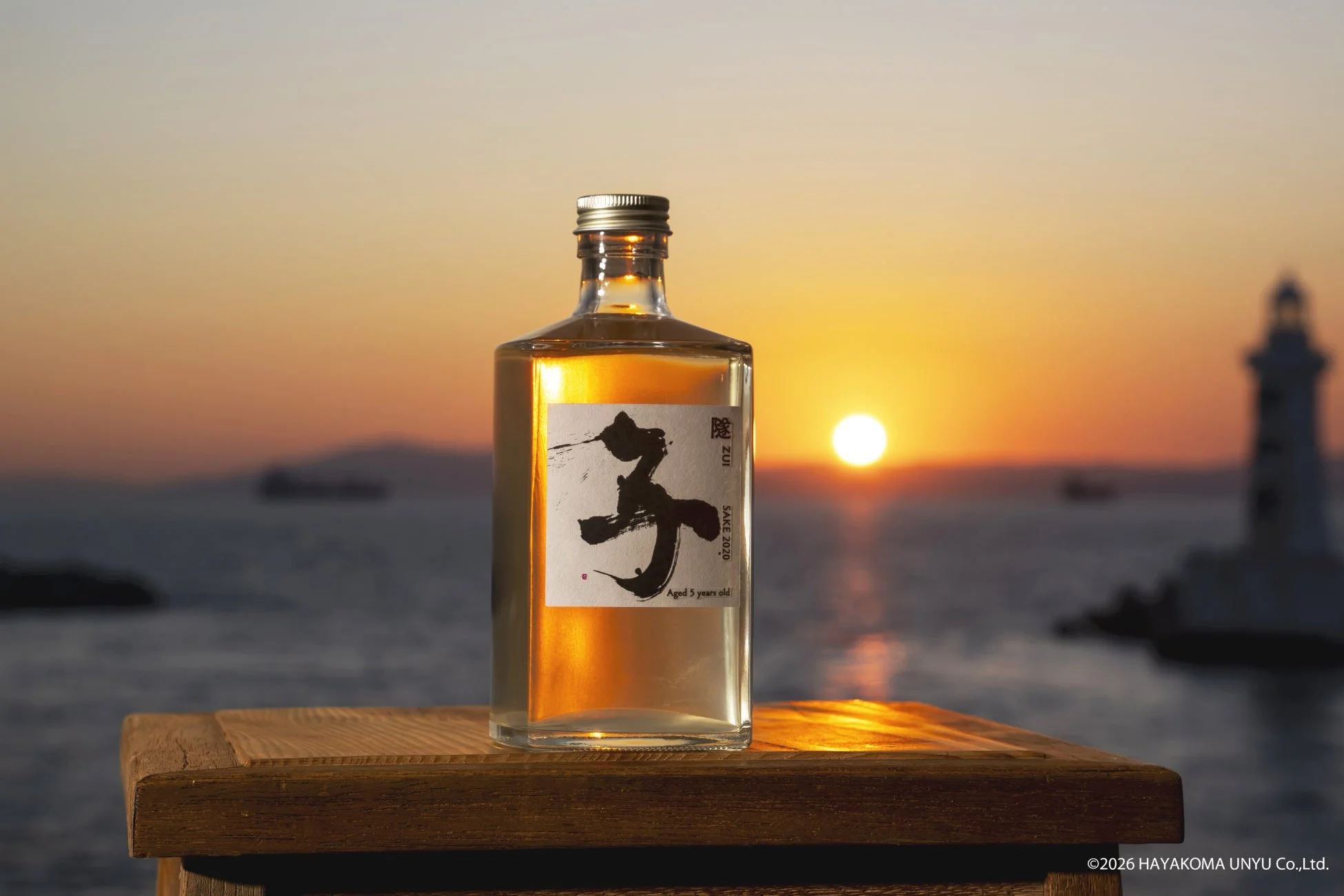 夕焼けの海を背景に、木製の台の上に置かれた日本酒のボトルが写っています。