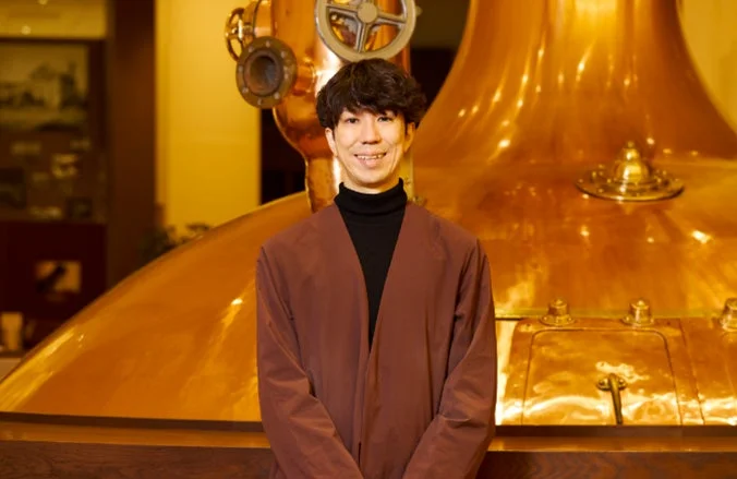 笑顔で醸造設備を背景に立つChief Experience Brewerの有友亮太氏