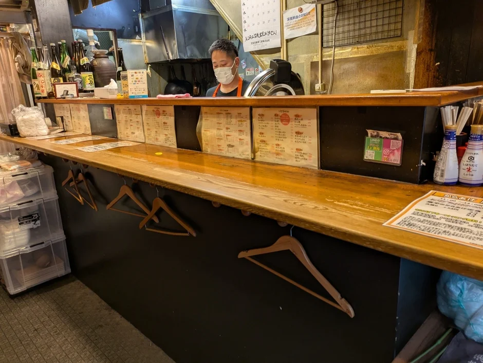 日本の居酒屋のカウンターの様子。マスクを着用した店員が立ち、カウンターにはメニュー、QRコード決済の案内、調味料が並んでいる。足元には衣類用のハンガーが見える。