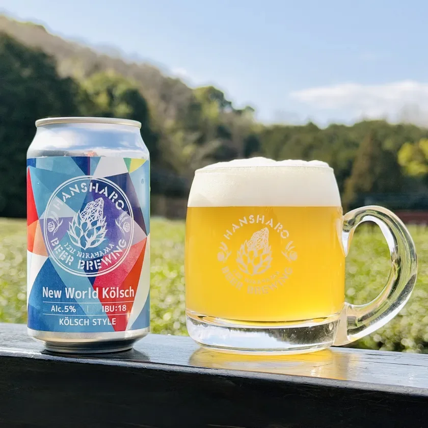 「HANSARO BEER BREWING」のクラフトビール「New World Kölsch」の缶と、同じビールが注がれたグラスが屋外に置かれています。背景には緑豊かな丘が広がり、晴れた日の爽やかな雰囲気が伝わってきます。