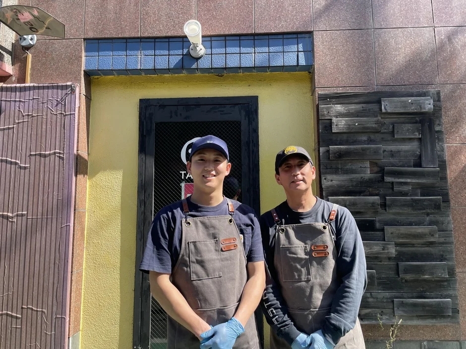 エプロンと帽子を着用した2人の男性が、黄色い店舗の前に立っています