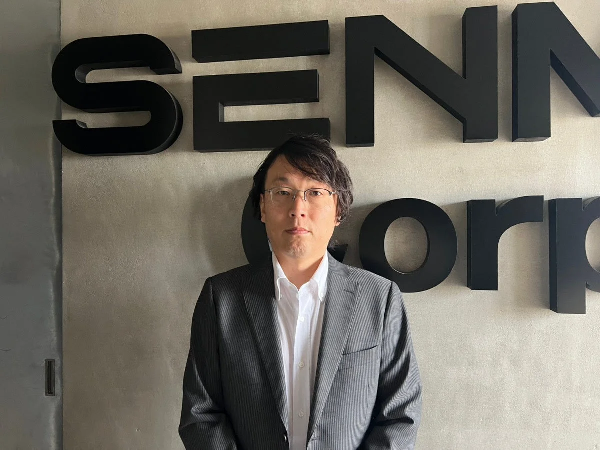 スーツを着た男性が、SENNA Corpのロゴが掲げられた壁の前で立っているビジネスシーンのポートレート