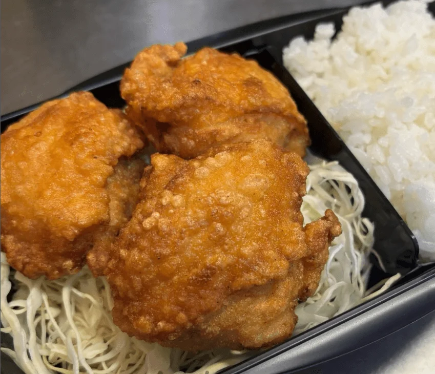黒い弁当箱に盛られた、揚げたての唐揚げ3個と白いご飯、千切りキャベツの食事です。美味しそうな和風のテイクアウト弁当の様子が写っています。