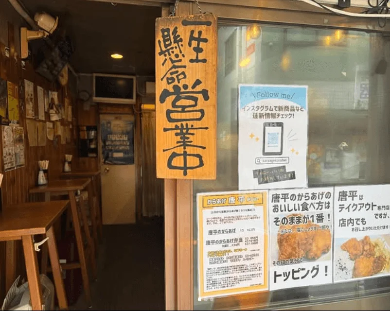 からあげ専門店「唐平」の店先と店内を捉えた写真。入口には「一生懸命営業中」の看板があり、窓にはインスタグラム案内やからあげのメニュー、食べ方に関するポスターが貼られている。木製のテーブルと椅子が並び、テイクアウトも店内飲食も可能なアットホームな店舗の様子。