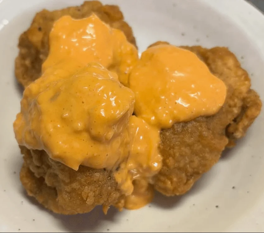 白い皿に盛られた、オレンジ色のクリーミーなソースがたっぷりとかかった揚げ物料理です。香ばしく揚がったチキンに濃厚なソースが絡み、食欲をそそる一品です。