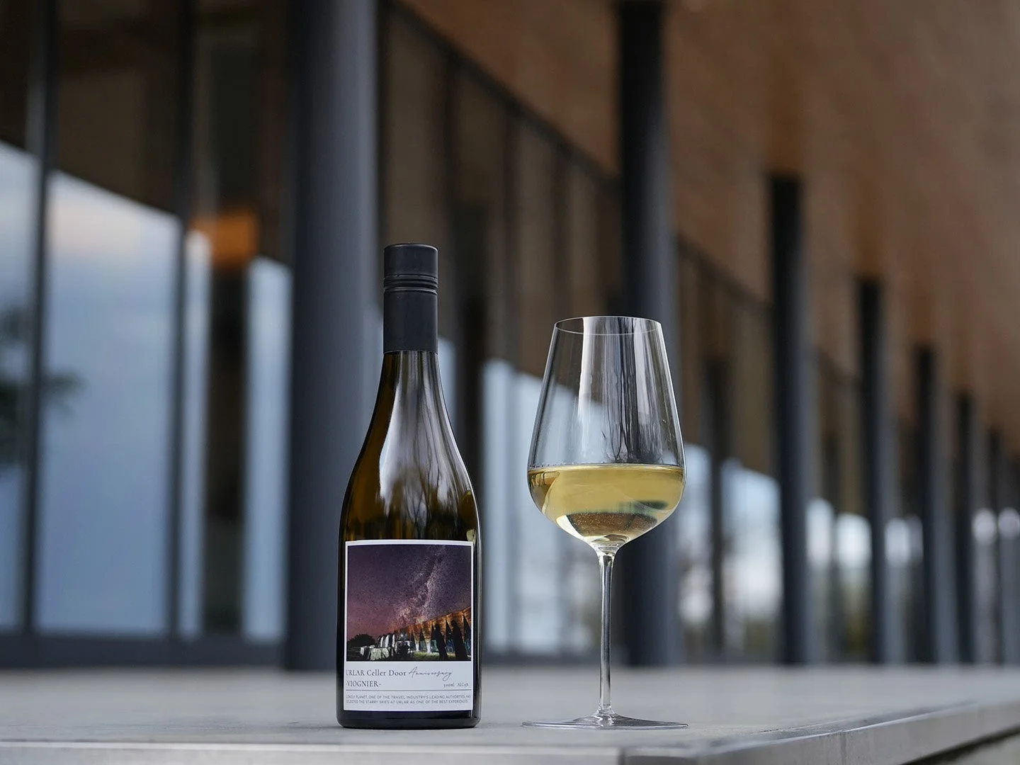 モダンな建物を背景に、白ワインのボトルとワイングラスが置かれています。ボトルには星空が描かれたラベルが貼られており、洗練された雰囲気です。
