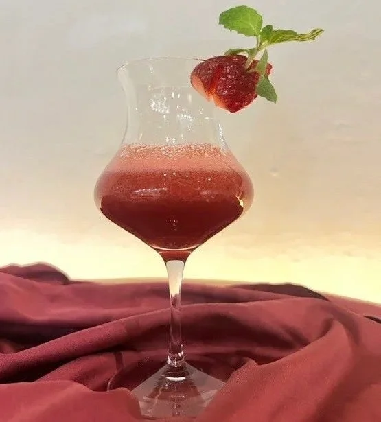 赤い飲み物が入ったグラスが、赤い布の上に置かれています。グラスの縁にはイチゴとミントの葉が飾られており、鮮やかで美味しそうなカクテルかスムージーのように見えます。美しい盛り付けが特徴です。