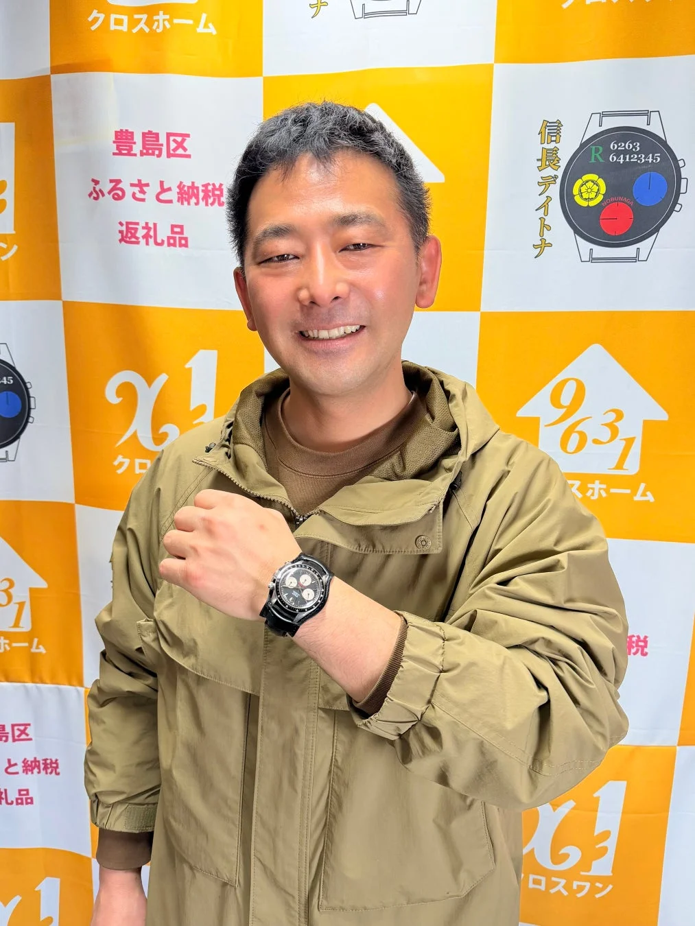 笑顔の男性がカーキ色のパーカーを着て腕時計を見せている画像。背景には「豊島区 ふるさと納税 返礼品」や「信長デイトナ」と書かれた黄色いチェック柄のバナーが見えます。