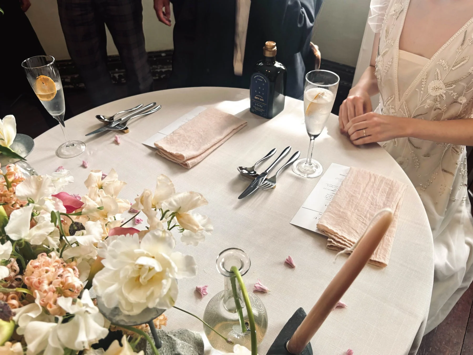 結婚披露宴と思われる会場のテーブルセッティングの様子を捉えた写真です。花嫁の姿、華やかなフラワーアレンジメント、カクテルグラス、ジンボトル、整えられた食器が写っており、上品で祝祭的な雰囲気が伝わります。