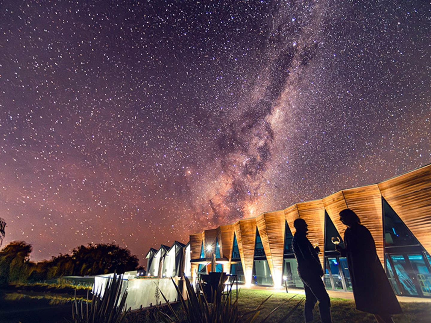 満点の星空の下、モダンな建物とシルエットになった二人の人物がワイングラスを傾けているロマンチックな夜景。天の川が印象的です。