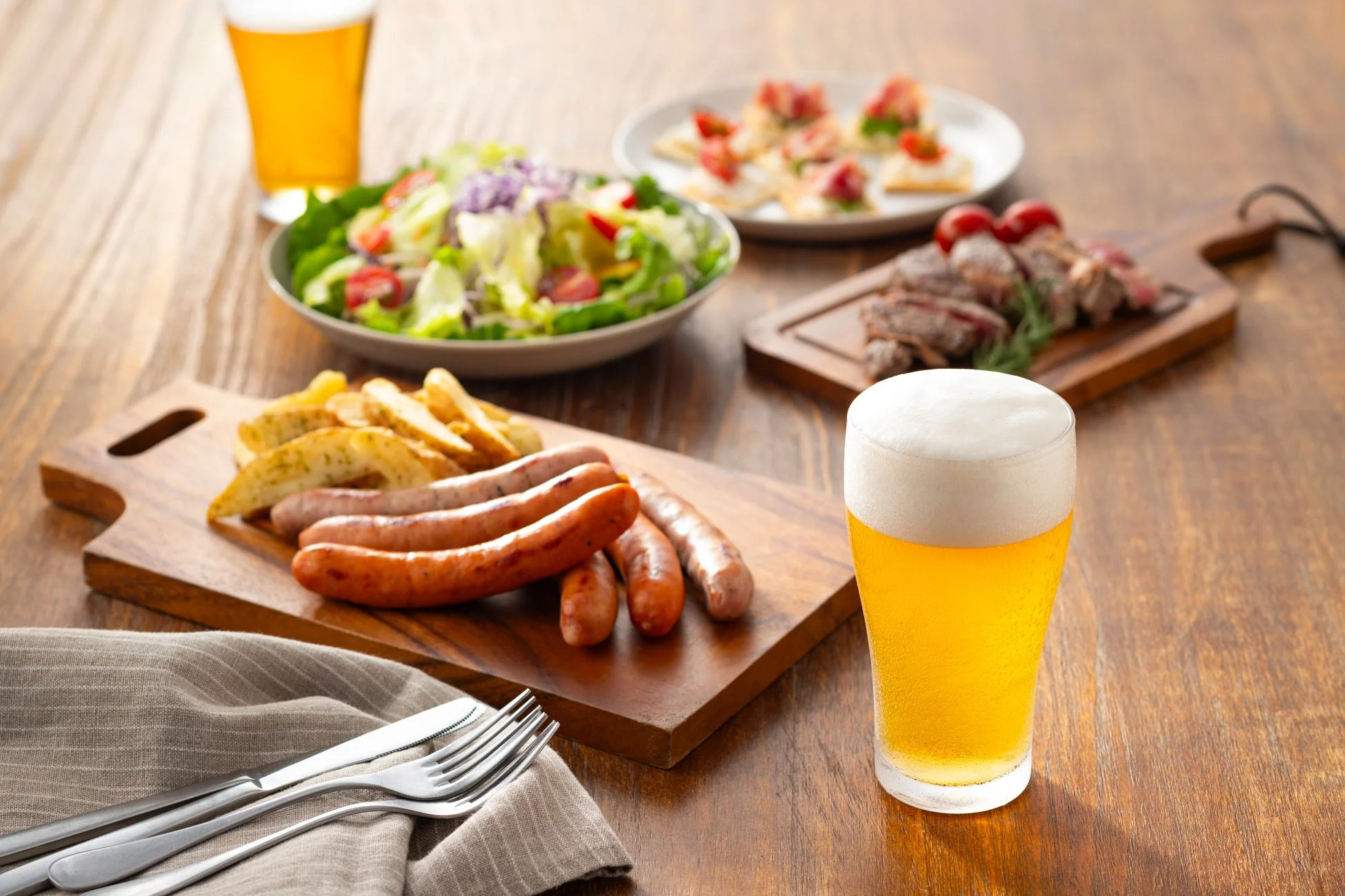 泡立つビールグラス、ソーセージとポテトの盛り合わせ、スライスされた肉料理、新鮮なサラダ、前菜が並べられた豪華な食事風景