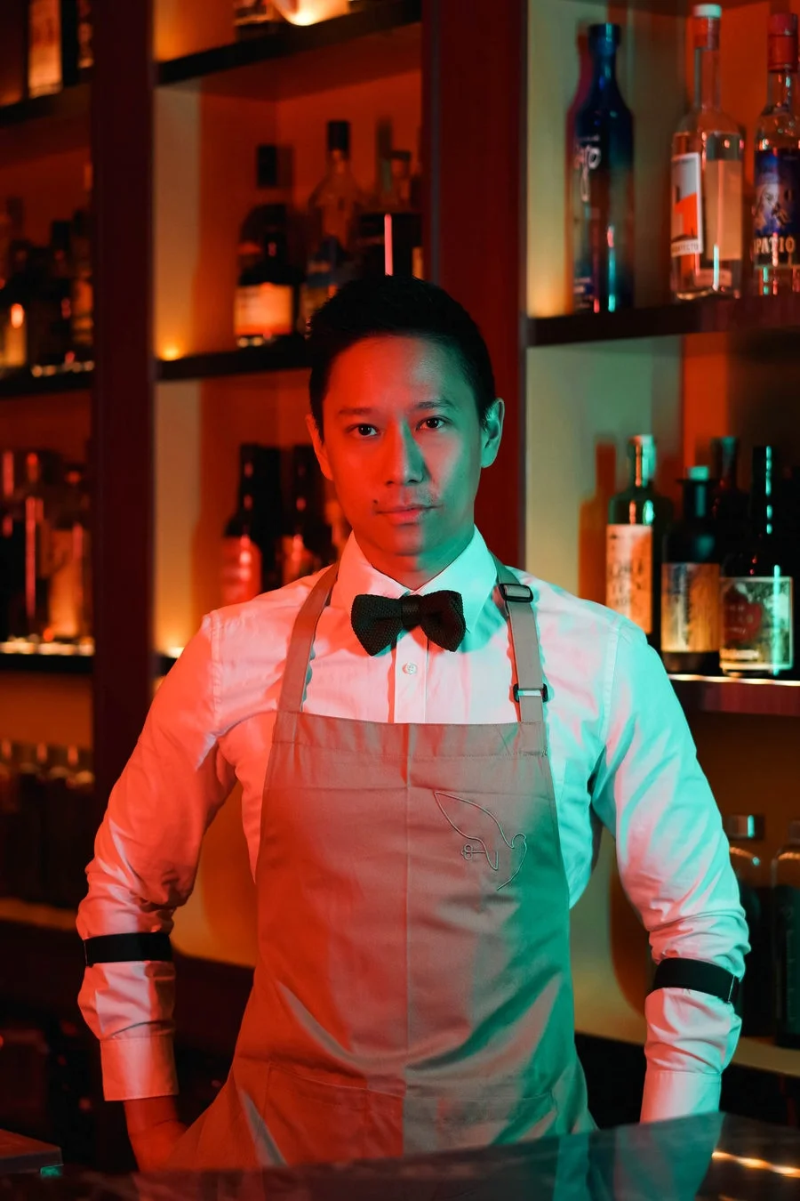 バーで立つ男性バーテンダーのポートレート。白シャツに蝶ネクタイとエプロンを着用し、赤と緑の照明に照らされています。背景には酒瓶が並んでいます。