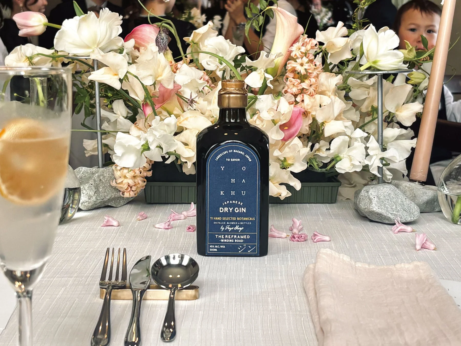 日本のドライジンボトルが、白とピンクの美しい花々で飾られたテーブルの主役です。グラスやカトラリーも整然と並び、優雅なイベントや食事の雰囲気が伝わります。背景には子供の姿も。
