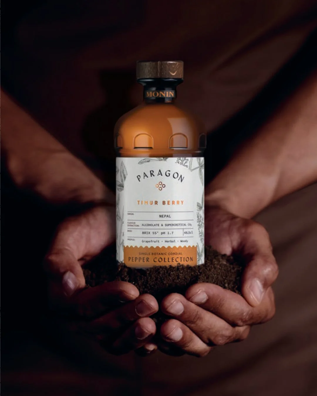 土をすくう両手に抱えられた「MONIN PARAGON TIMUR BERRY」のボトルが写っています。ネパール産のティムールベリーを特徴とし、グレープフルーツ、ハーブ、ウッディな香りが楽しめるシングルボタニカルコーディアルです。