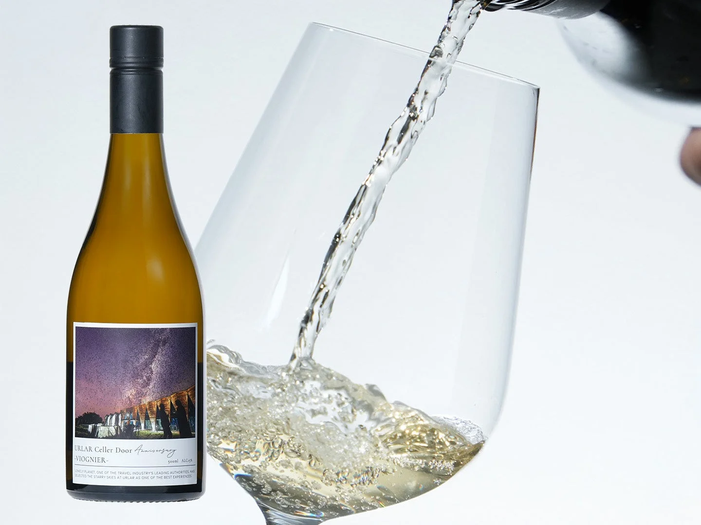 白い背景に、ワインがグラスに勢いよく注がれている瞬間と、独特なラベルデザインの白ワインボトルが写っています。ボトルのラベルには星空と建物が描かれており、特別な体験を想起させます。