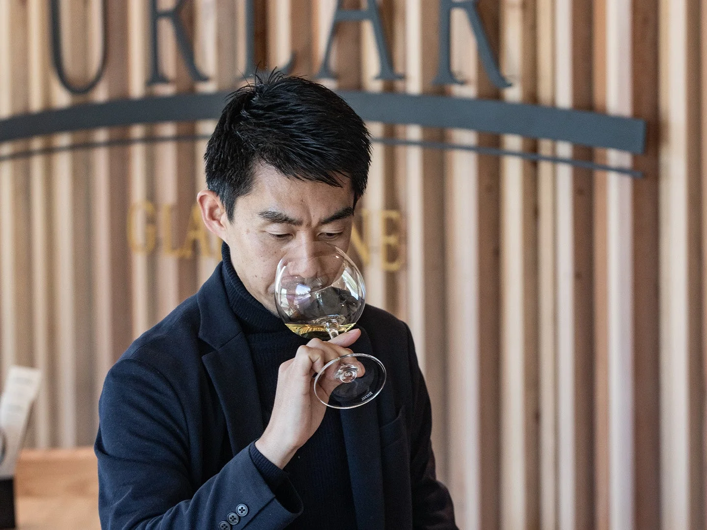 一人の男性が真剣な表情で白ワインの香りを嗅いでいる様子を捉えた画像です。彼は黒いタートルネックとジャケットを着用し、ワイングラスを手にしています。背景には木目調の壁と「URLAR」という文字が見え、ワイナリーや試飲会場のような雰囲気です。