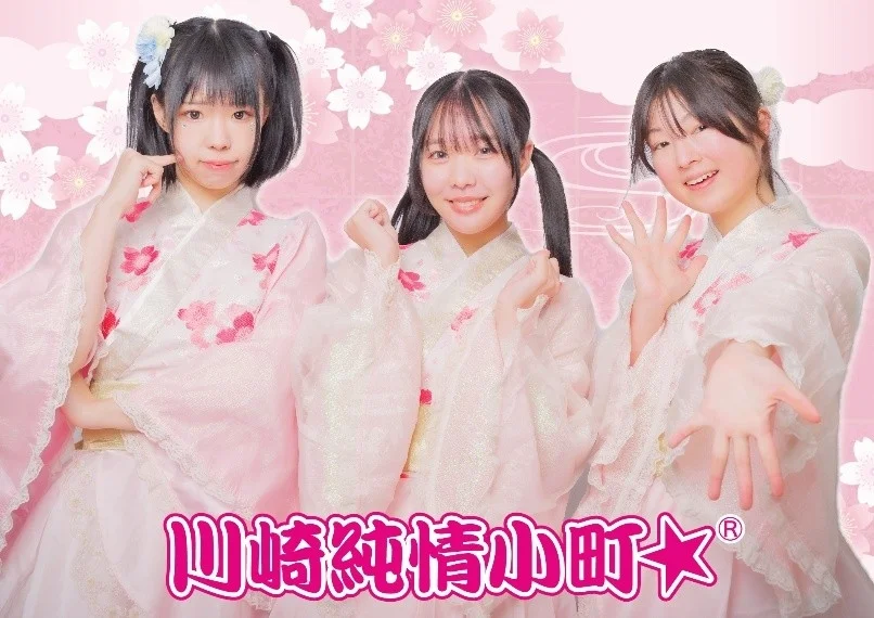 桜柄のピンクの衣装を着た3人の女性アイドルグループ「川崎純情小町☆」が、笑顔でポーズをとっている
