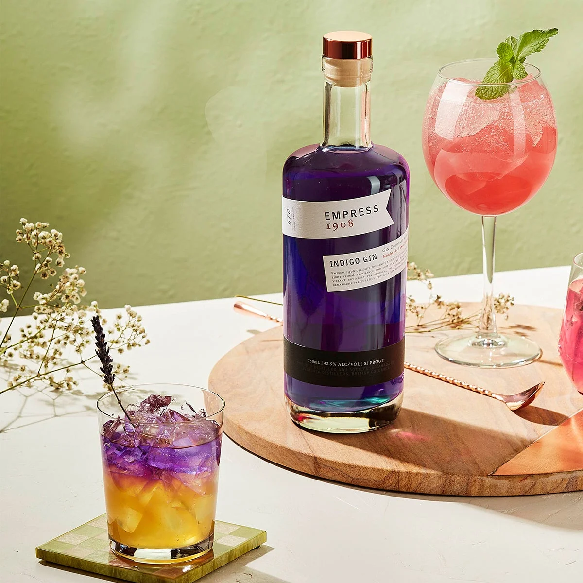 紫色の「Empress 1908 Indigo Gin」のボトルと、それを使った色鮮やかな2種類のカクテルが並べられています。手前のカクテルは黄色と紫のグラデーションでラベンダーが添えられ、奥の赤いカクテルにはミントが飾られており、おしゃれで涼しげな雰囲気を醸し出しています。