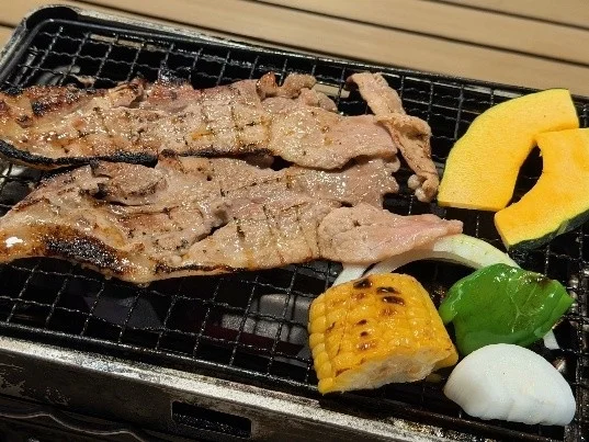 網の上で焼かれている肉と様々な野菜（カボチャ、トウモロコシ、玉ねぎ、ピーマン、餅）が写っており、食欲をそそるバーベキューの光景