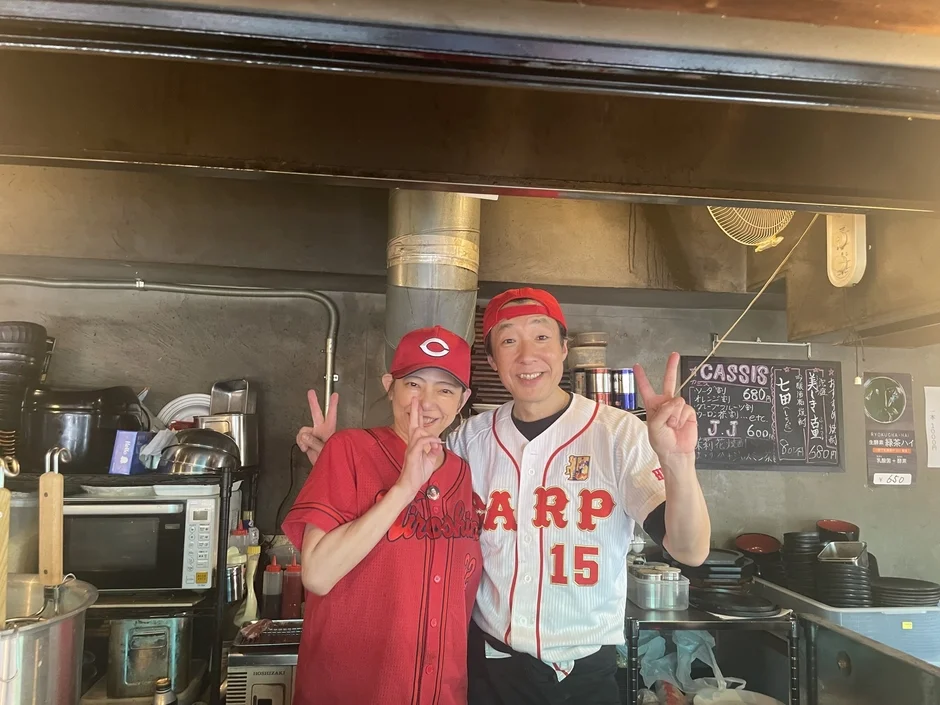 広島東洋カープのユニフォームとキャップを着用した男女が、飲食店のカウンターで笑顔でピースサイン
