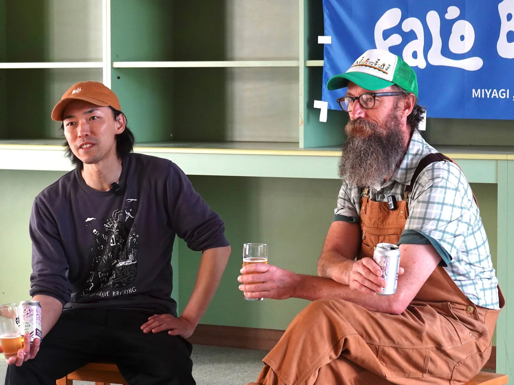 二人の男性が屋内でビールを飲みながら座っている。キャップをかぶった日本人男性と、長いひげの外国人男性がカジュアルな服装で、ビール関連のイベントで交流している様子。背景には「Falo B MIYAGI」のバナーが見える。