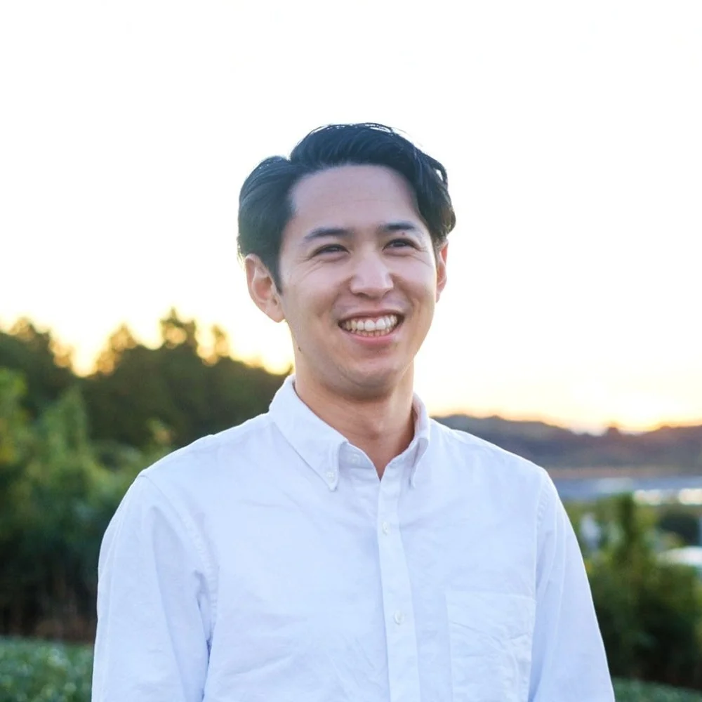 白いシャツを着たアジア人男性が屋外で明るく微笑んでいます。背景には夕焼けのような優しい光と木々が見え、爽やかな印象のポートレートです。
