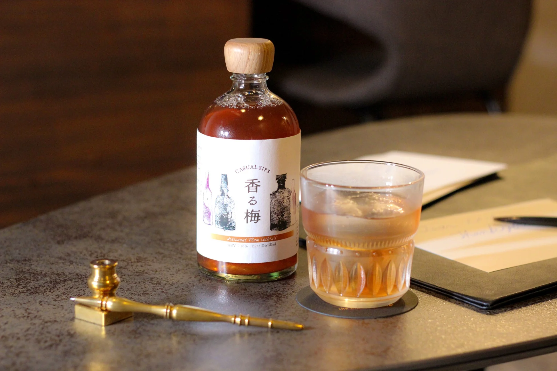 テーブルの上に「CASUAL SIPS 香る梅 Artisanal Plum Cocktail」のボトルと、氷の入ったグラスが置かれています。