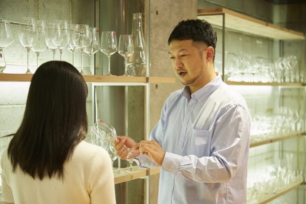 男性が女性にワイングラスを手に取りながら説明している様子です。背景には多くのワイングラスが並べられた棚があり、ワイングラスの専門店かショールームでの一場面と思われます。