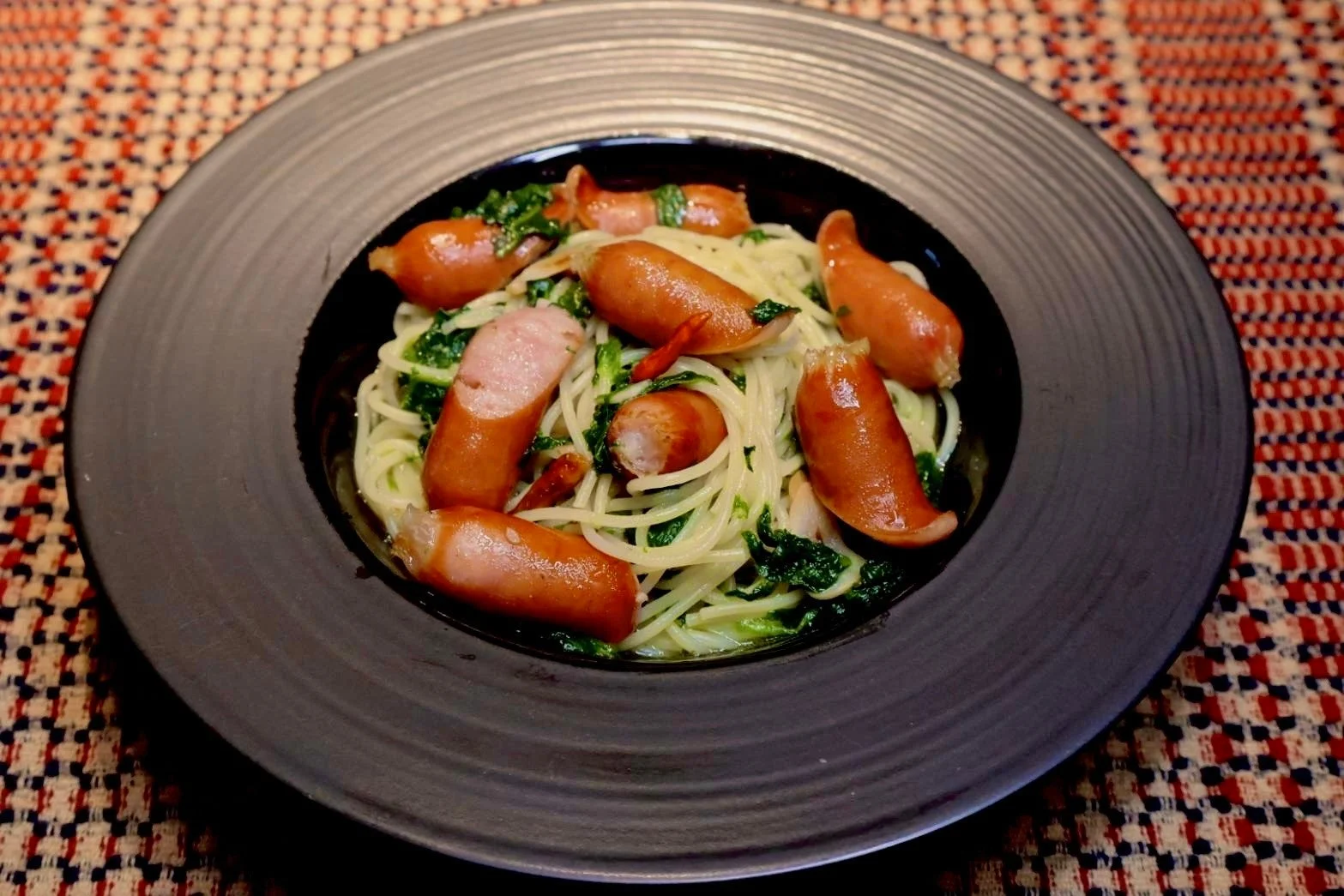 黒い皿に盛られたソーセージと緑の野菜が特徴のパスタ料理