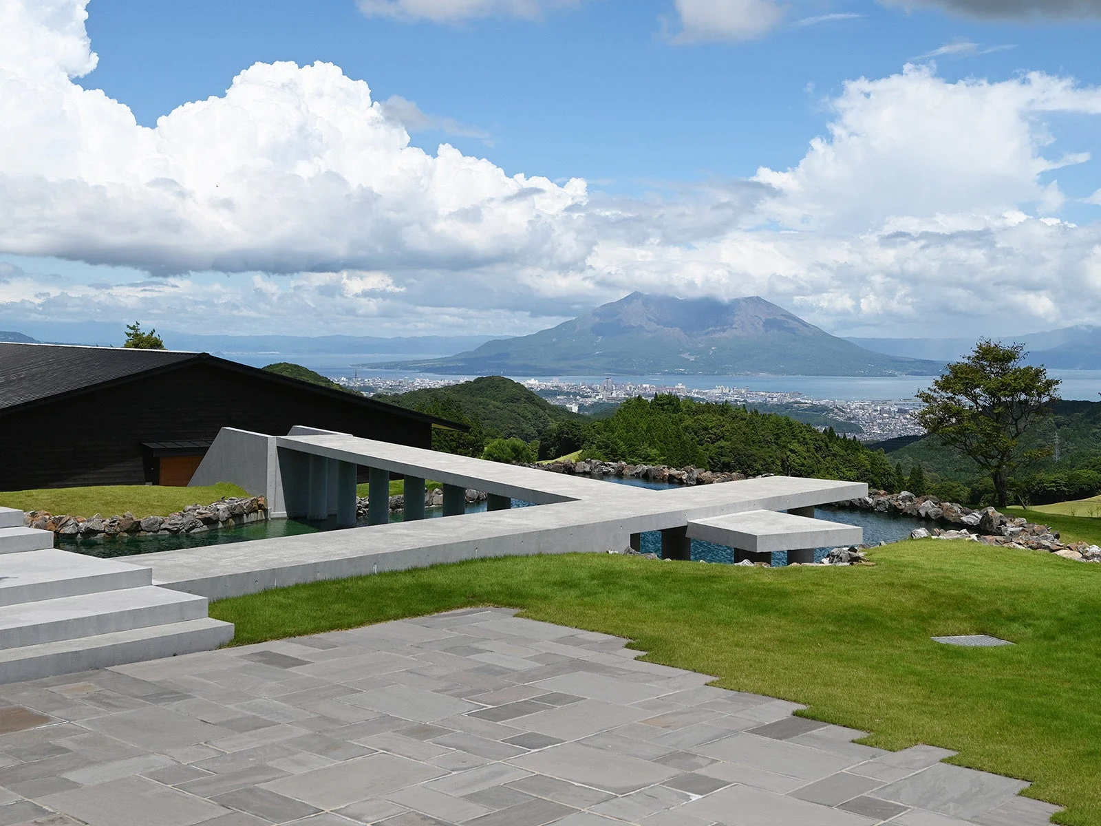 モダンなコンクリート構造と水辺の庭園、奥には桜島と鹿児島市街が望める壮大な風景
