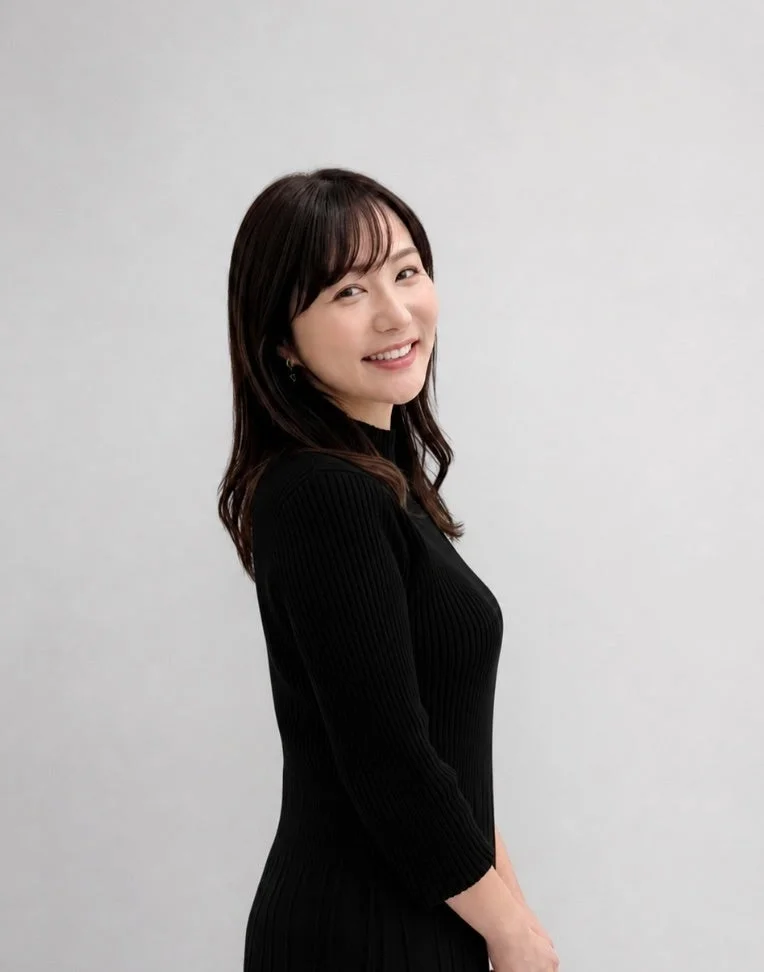 白い背景の前で、黒いリブ編みの服を着た女性が、カメラに向かって優しく微笑んでいます。