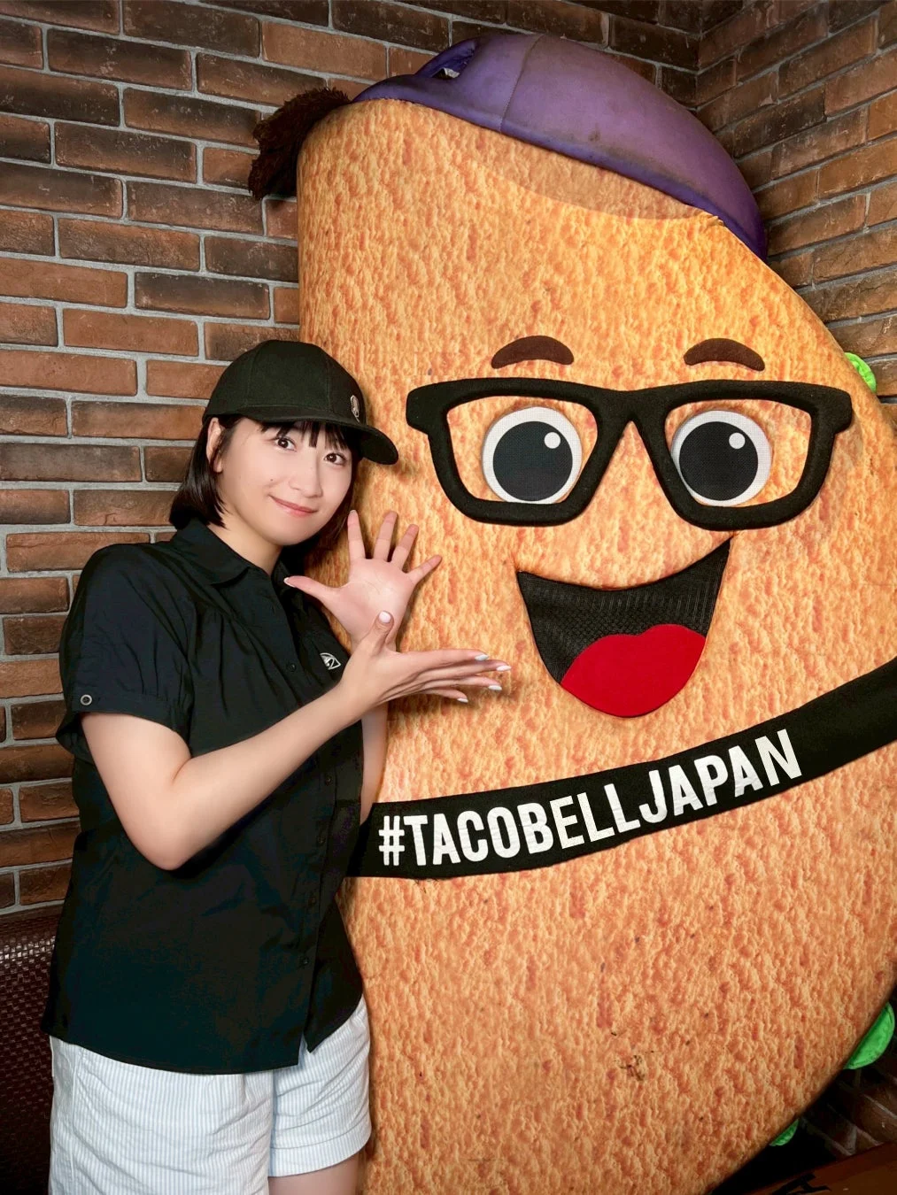 女性がタコベルの大きなタコ型マスコットの隣で笑顔でポーズをとっています。マスコットには「#TACOBELLJAPAN」と書かれており、背景はレンガの壁です。