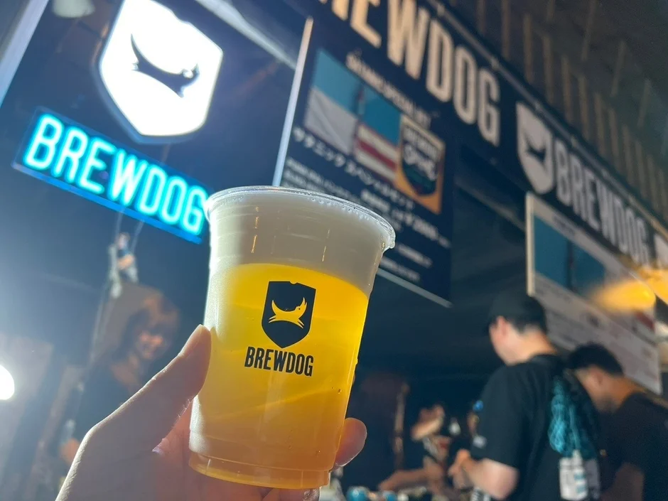 BREWDOGのネオンサインが光る場所で、ロゴ入りのカップに入ったビールを手にしている様子