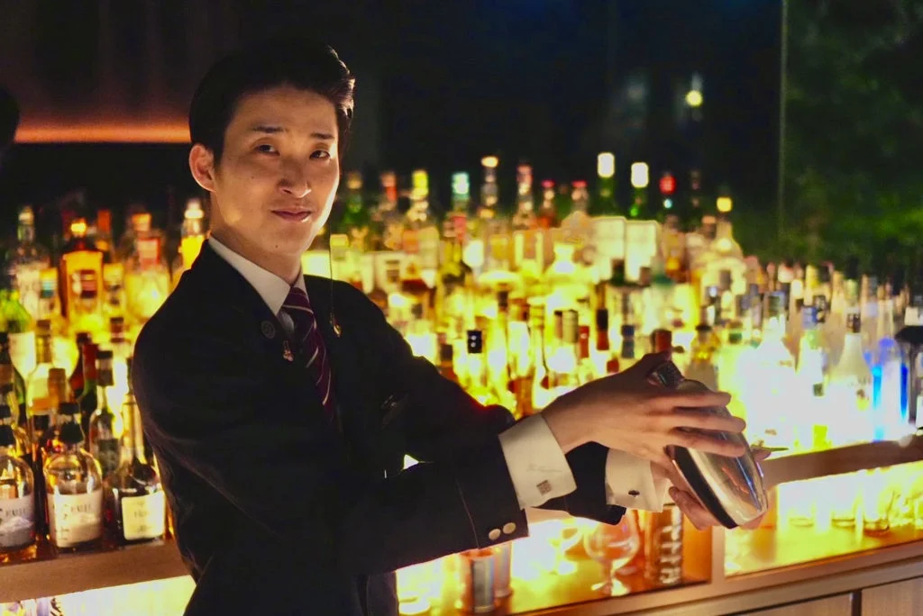 スーツを着た男性バーテンダーが、薄暗いバーカウンターでカクテルシェーカーを振っています。