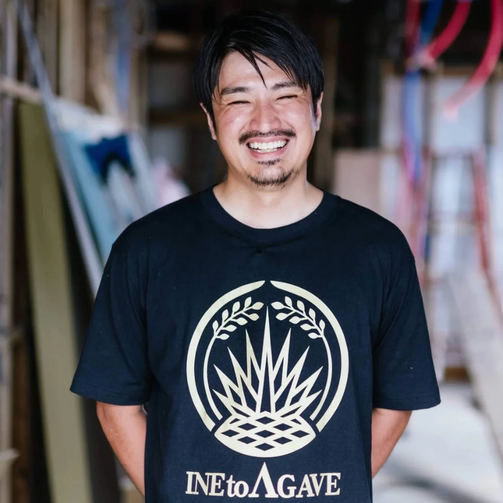 黒いTシャツを着て笑顔を見せる男性のポートレート。Tシャツには「INE to AGAVE」の文字と特徴的なアガベのようなロゴがデザインされています。背景はぼかされており、屋内であることが伺えます。
