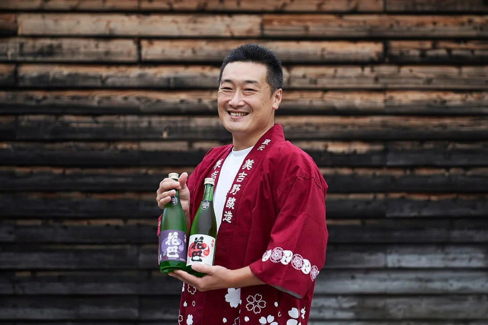 赤い法被を着て日本酒「花巴」のボトルを持つ美吉野醸造の橋本晃明氏