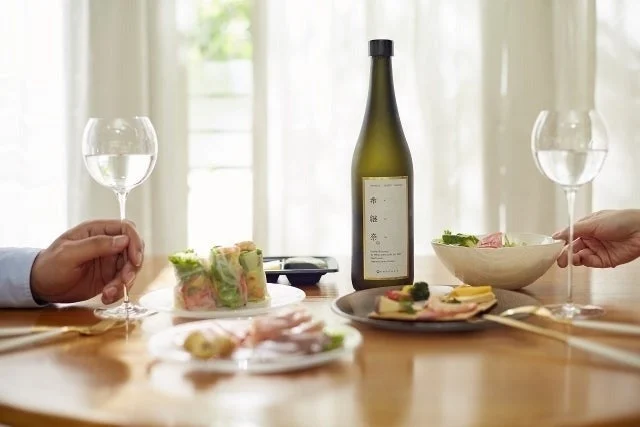 明るいダイニングで、日本酒のボトルとグラス、そして様々な料理が並んだ食卓を囲む二人の手が写っています。和やかな食事のひとときを表現しています。