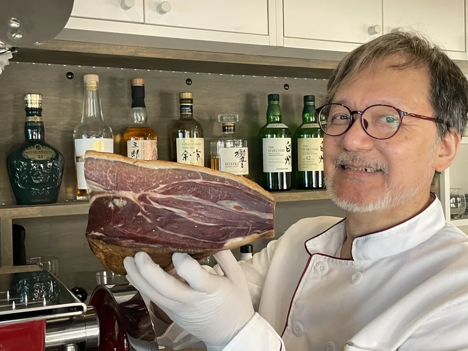 シェフの白衣を着た男性が笑顔で大きな生ハムの塊を持っている