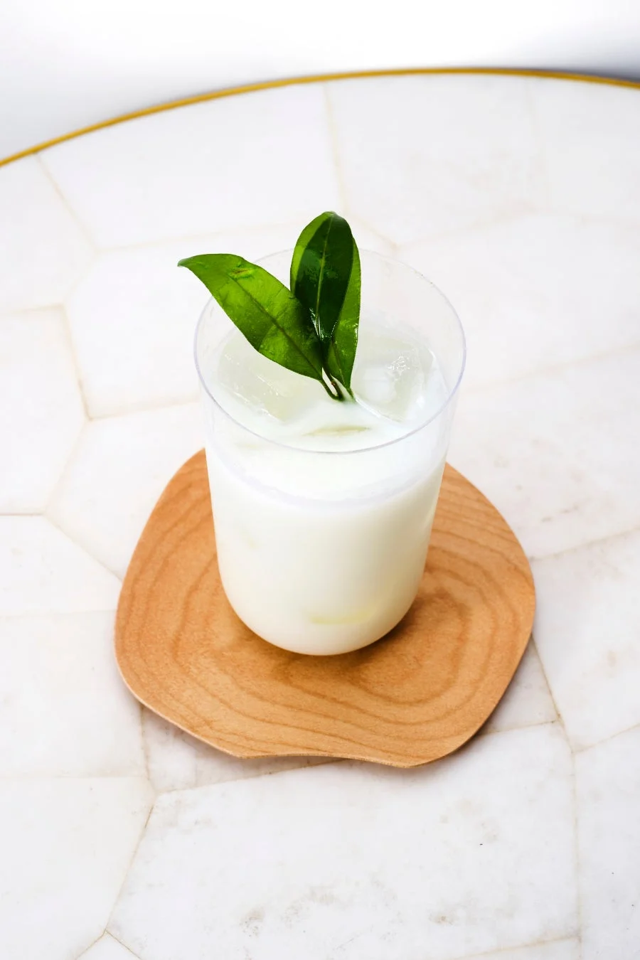 白い飲み物が入ったグラスが木製のコースターに乗せられており、二枚の緑の葉が飾られています。白い大理石のようなテーブルの上で、清潔感があり涼しげな印象の画像です。