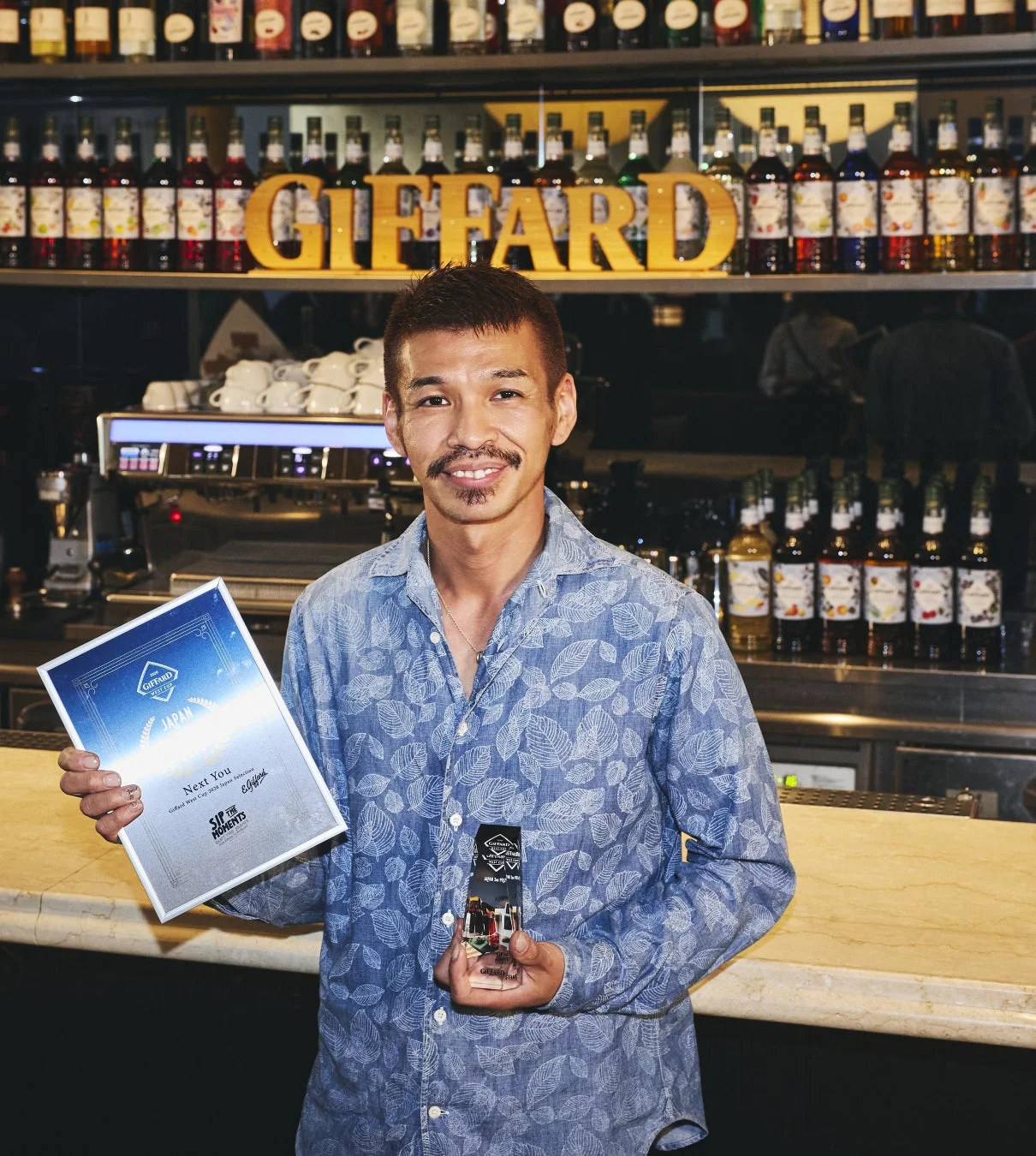 Giffard West Cup 2020 Japan Selectionの受賞者である男性が、バーカウンターを背景に笑顔で表彰状とトロフィーを手にしている。棚にはGiffardのボトルが多数並んでいる。