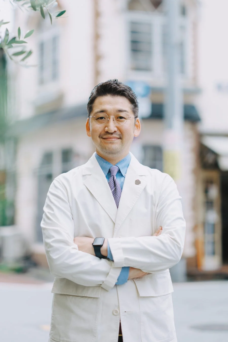 白衣を着て眼鏡をかけたアジア人男性が、腕を組んで笑顔で立っているポートレート写真です。