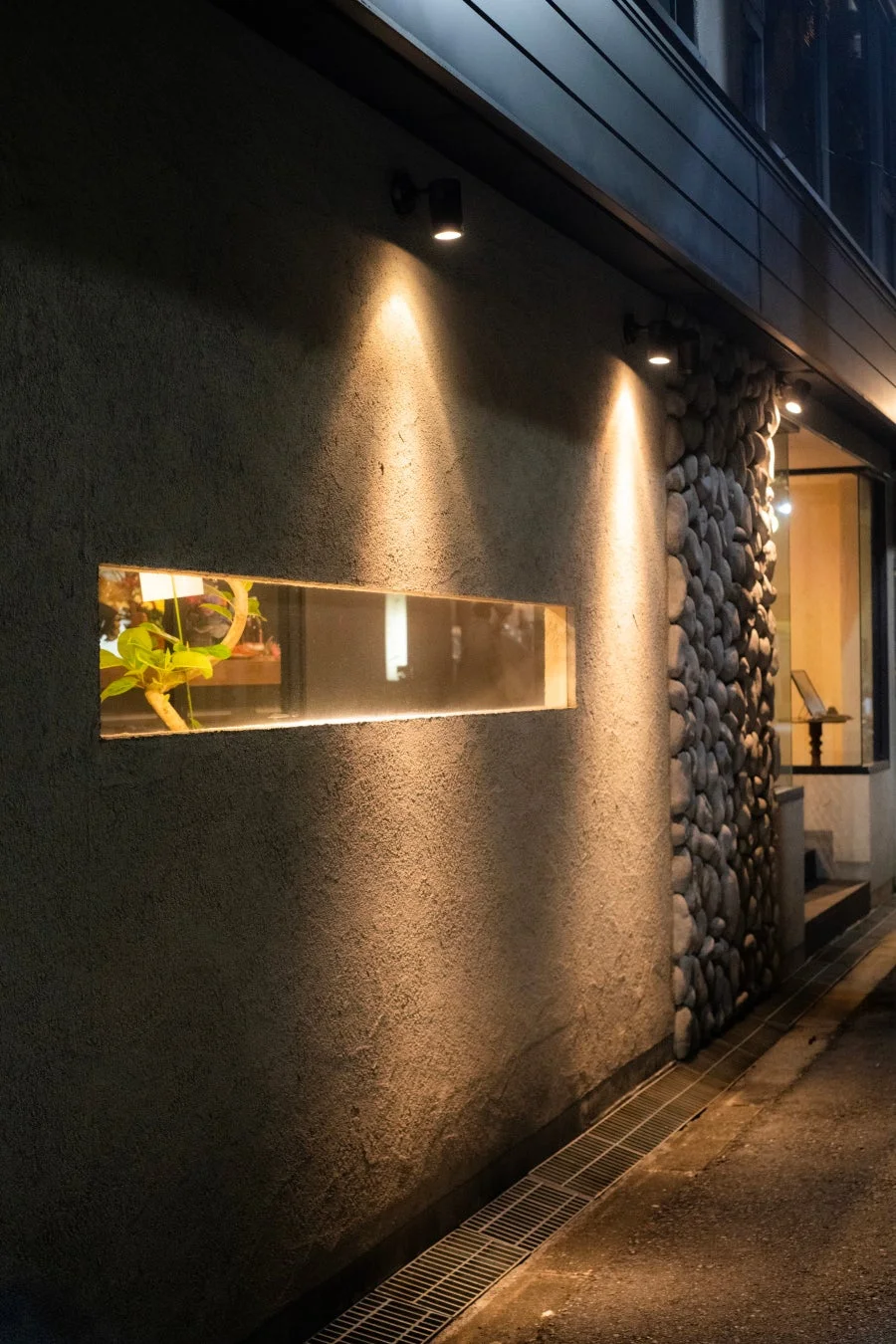 夜の建物の外壁が暖かなスポットライトで照らされています。壁には横長のスリット窓があり、中に植物が見えます。
