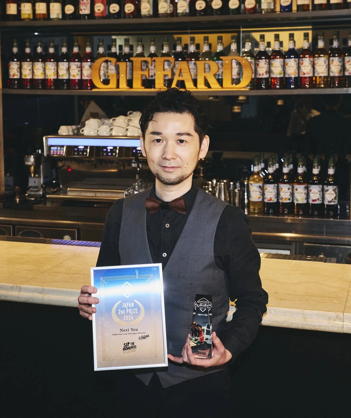 バーカウンターの前で、Giffardのロゴを背景に、蝶ネクタイとベストを着用した男性がGiffard West Cup 2026 Japan Selectionの2位の賞状とトロフィーを持っている様子です。