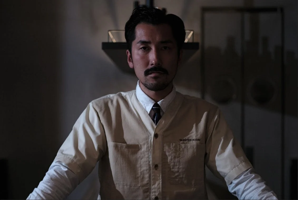 「QUARTER ROOM」と書かれたジャケットを着用した男性が、薄暗い室内でカメラをじっと見つめているポートレート。