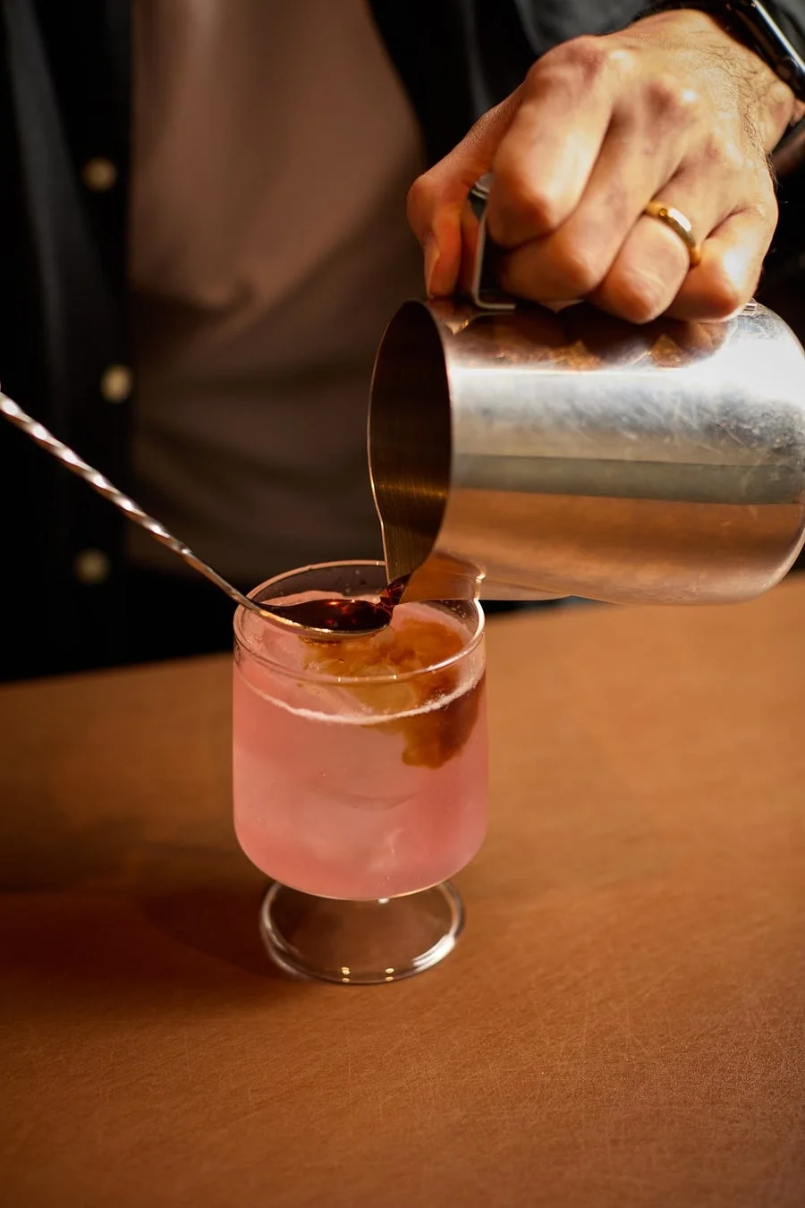 ピンク色の飲み物が入ったグラスに、手元が金属製のピッチャーから濃い色の液体を注ぎ入れている様子。