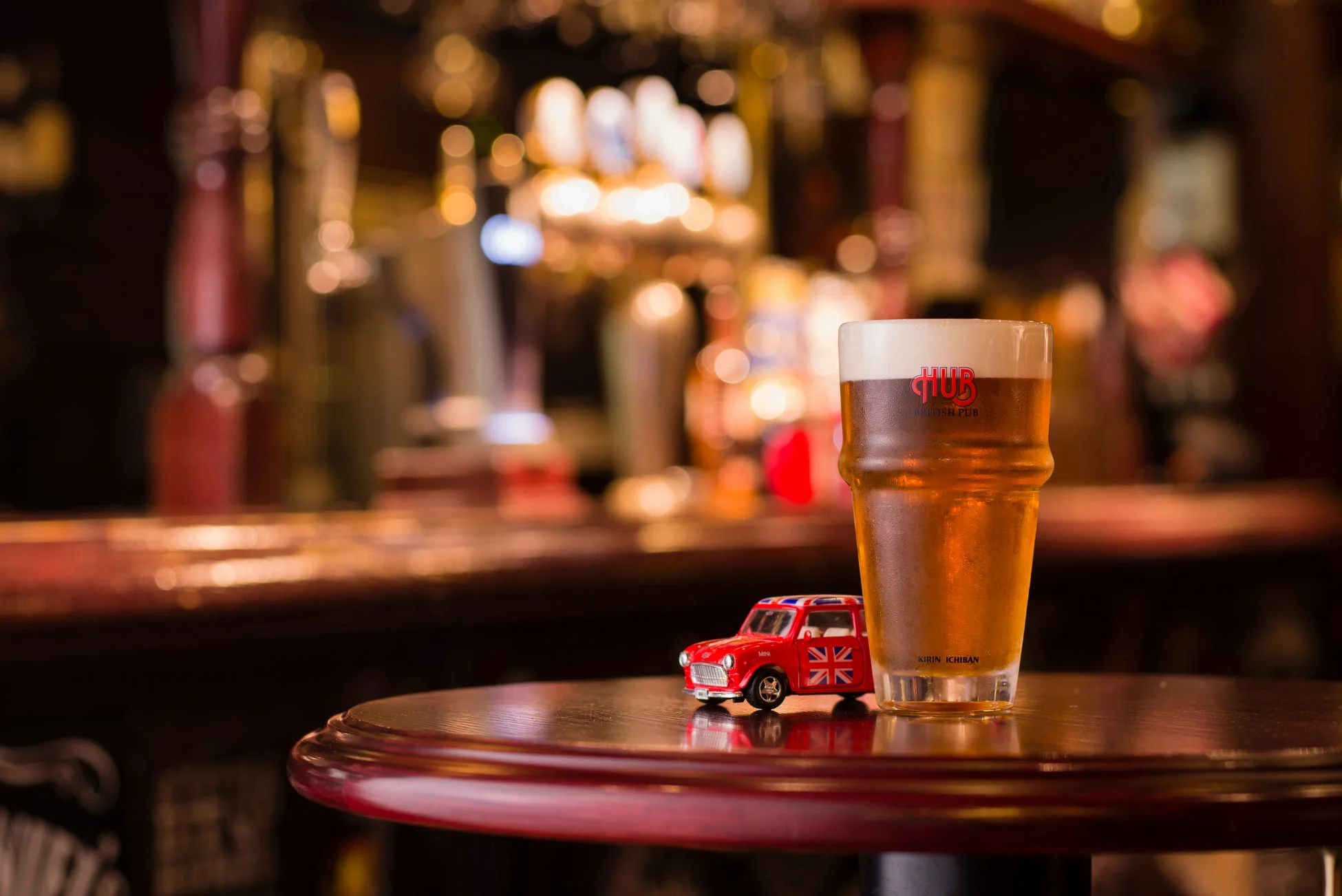 英国風パブのテーブルに、HUBのロゴが入ったビールグラスとユニオンジャック柄の赤いミニカー