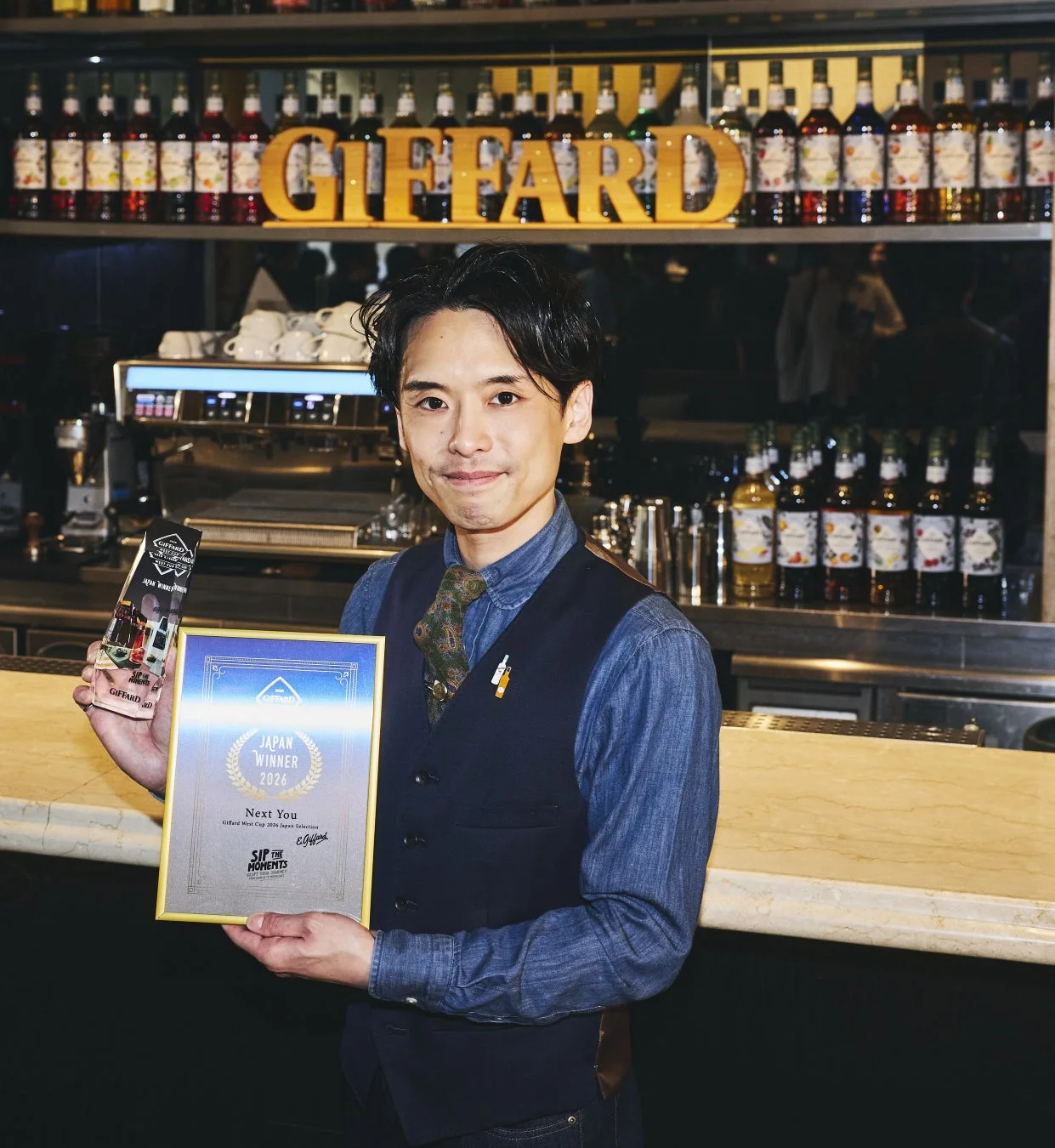 バーテンダーが「Giffard West Cup 2024 Japan Selection」の優勝賞状とトロフィーを手に笑顔で立っています。背景にはGiffardのシロップが並び、バーカウンターの雰囲気が伝わります。